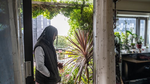One of the judges of the National Trust's Sky Gardening Challenge 2025 stands in the doorway to a balcony which is covered in lush green plants. Beyond is a view of the Manchester skyline.